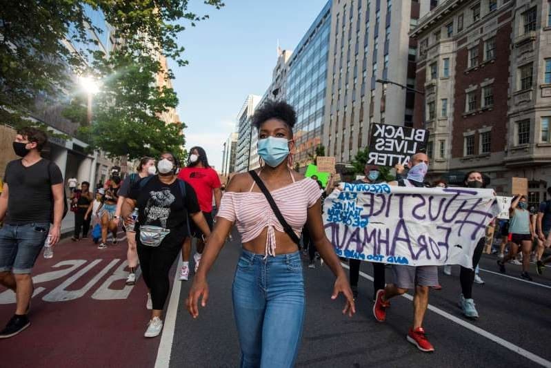 Color photo of a young Black woman marching with a face mask. There are other marchers behind her on a city street. Visible signs include BLACK LIVES MATTER and #JusticeForAhmaud.