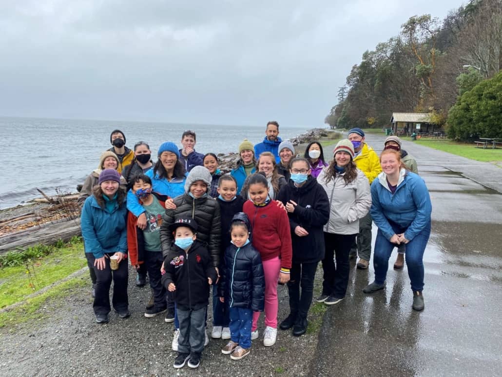 A group of TeamChild current and alumni staff and family members are gathered by the seashore on a grey day. Many wear hats and several wear face masks.