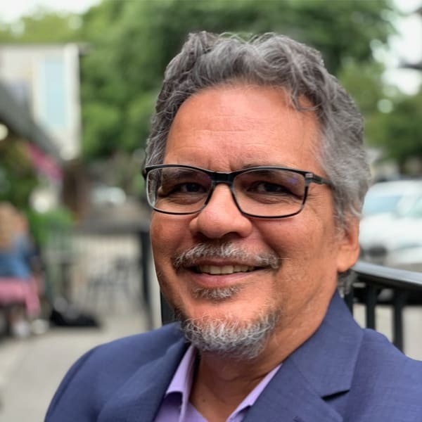 TeamChild Executive Director Marcos Martinez is pictured outside. He wears glasses and a smokey blue blazer and smiles into the camera.