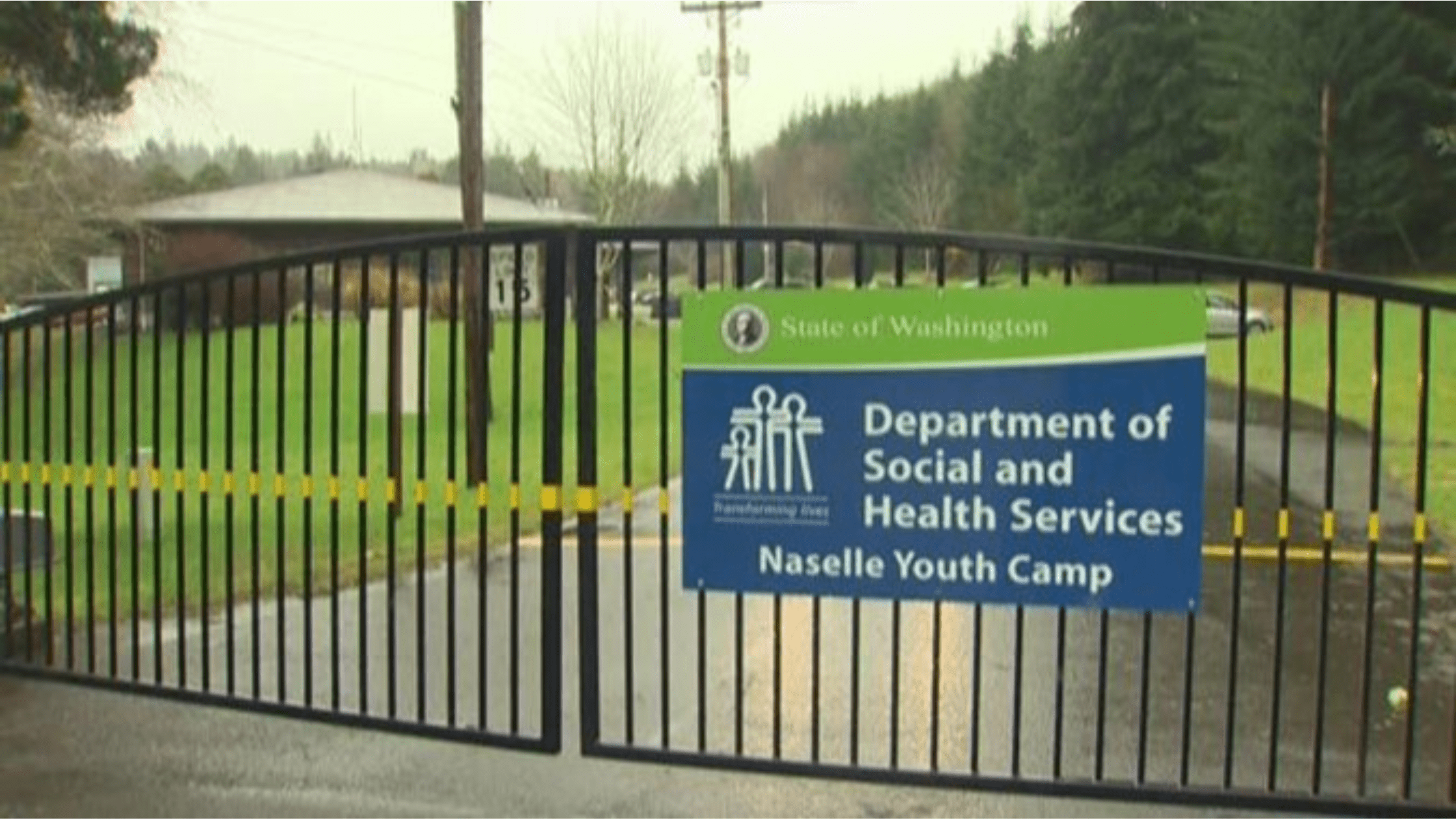 A building and lawn are pictured behind a closed gate. The sign on the gate reads: State of Washington - Department of Social and Health Services - Naselle Youth Camp.