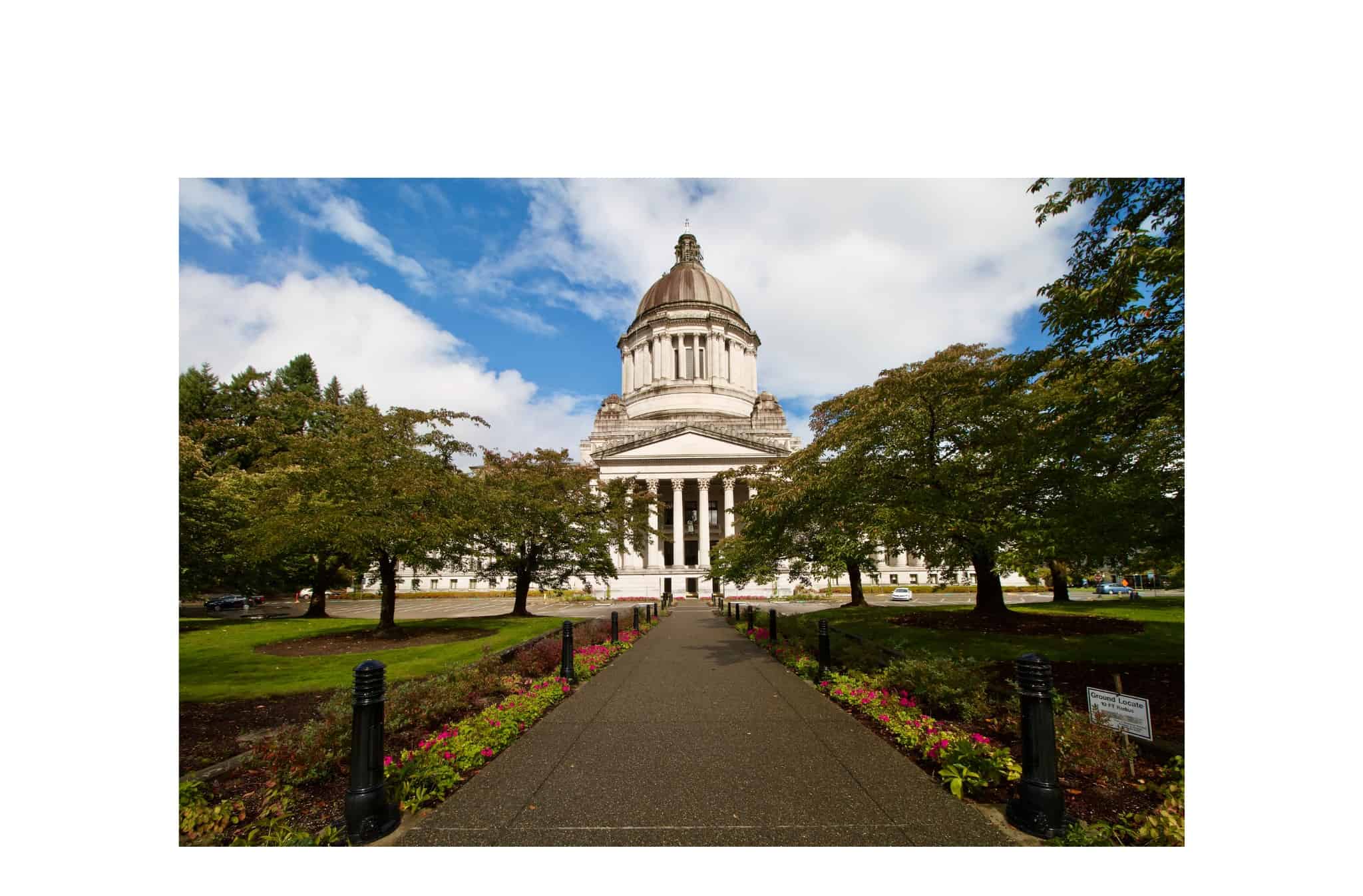 The State Capitol buiding in Olympia is pictured against a blue sky with clouds - there are trees and a walkway with flowers in the foreground.