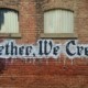 A brick wall with three shuttered windows features a mural and painted text reading: Together We Create!