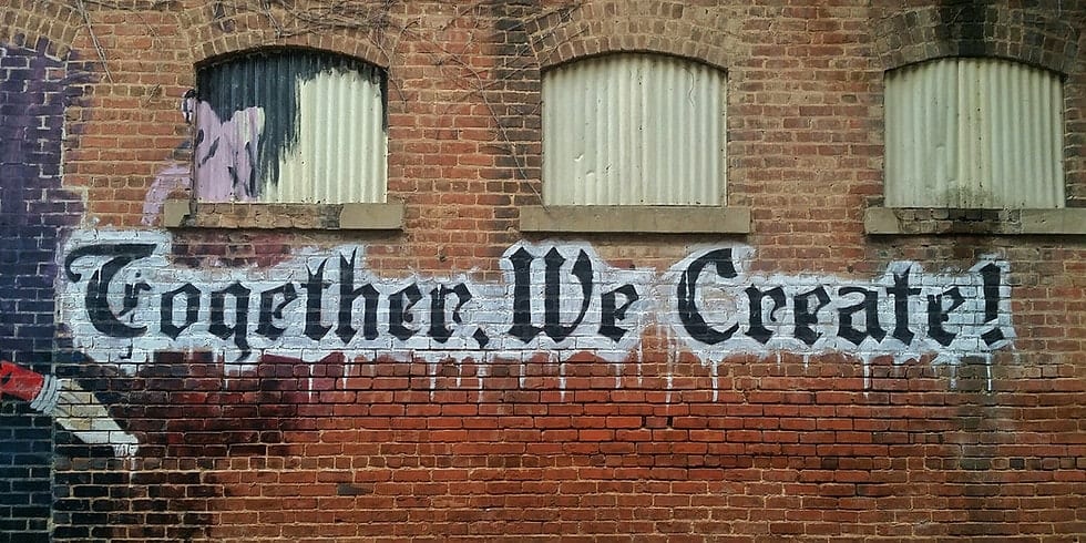 A brick wall with three shuttered windows features a mural and painted text reading: Together We Create!