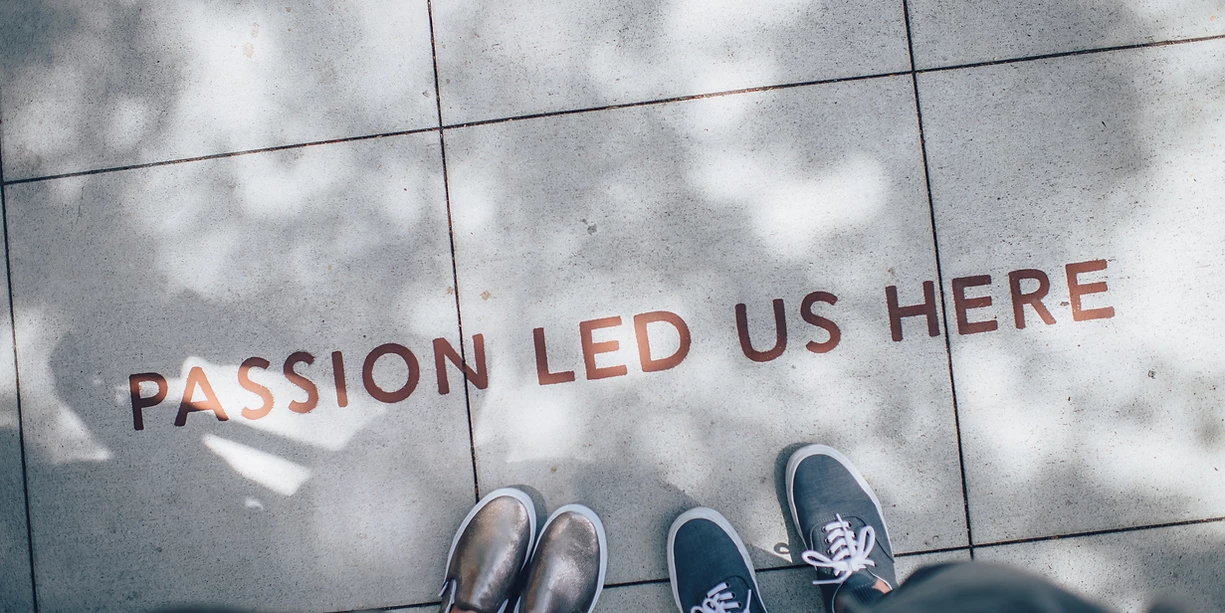 Two sets of shoes are pictured on a sidewalk with filtered light. Text reads: Passion Led Us Here