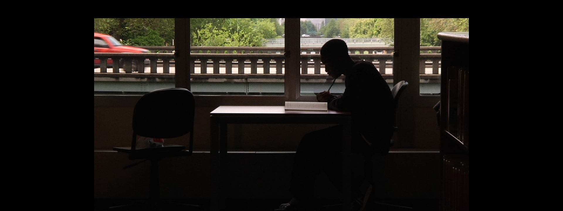 A young person is working at a desk. A red car is crossing a bridge in the background.