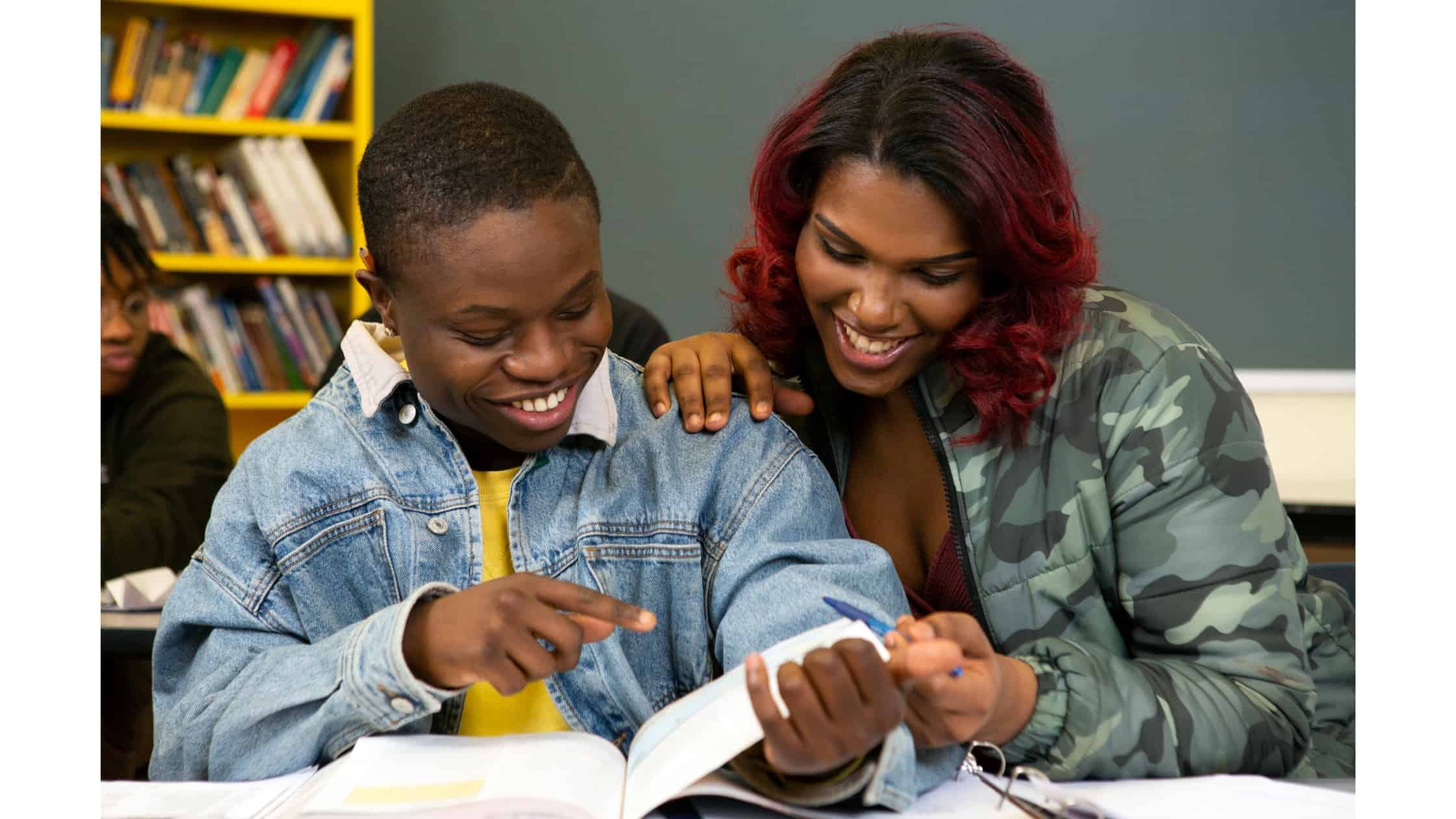 An adult and a young person look at a book together at a desk. They are smiling and another student sits behind them in the classroom.