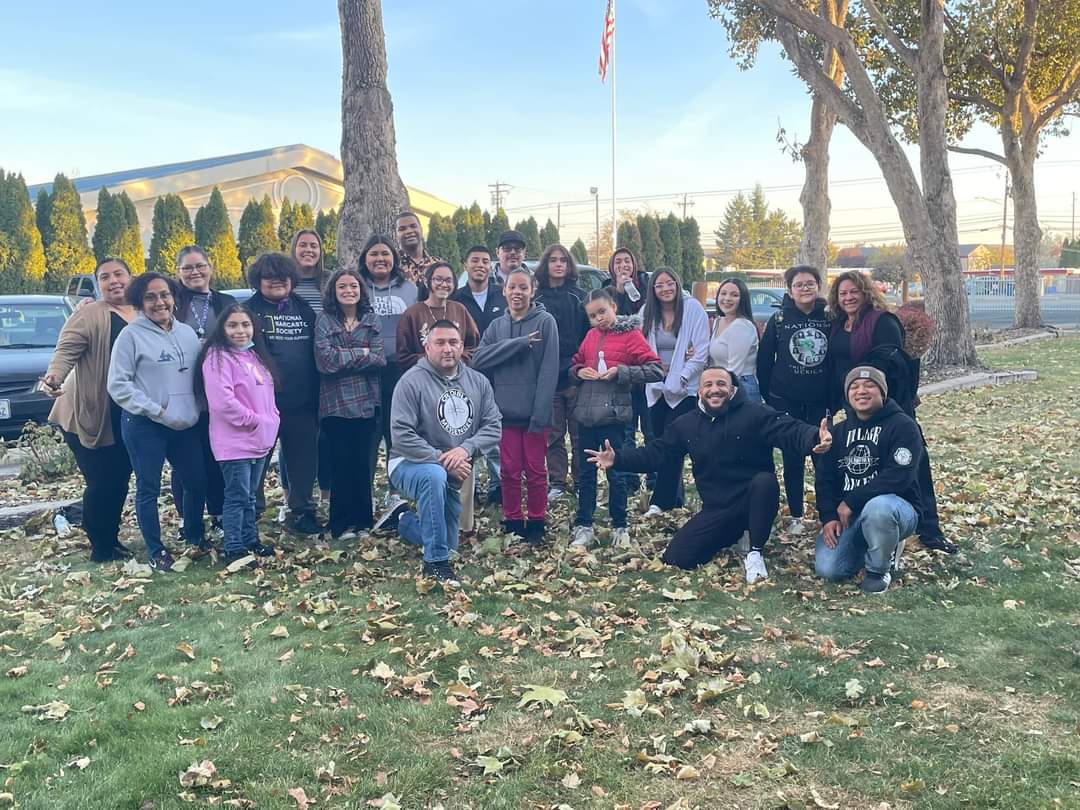 A group of young people are gathered on a lawn with fall leaves around them. There is a flag pole and some trees in the background. TeamChild staff members Porsche, Sonia and Quincy are among those pictured.
