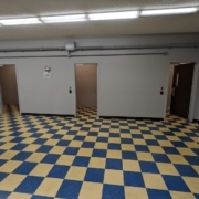 A checkered floor is shown beneath three doors that lead to small windowless rooms. There are fluorescent lights above.