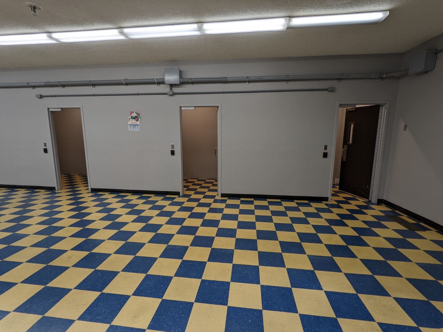 A checkered floor is shown beneath three doors that lead to small windowless rooms. There are fluorescent lights above.
