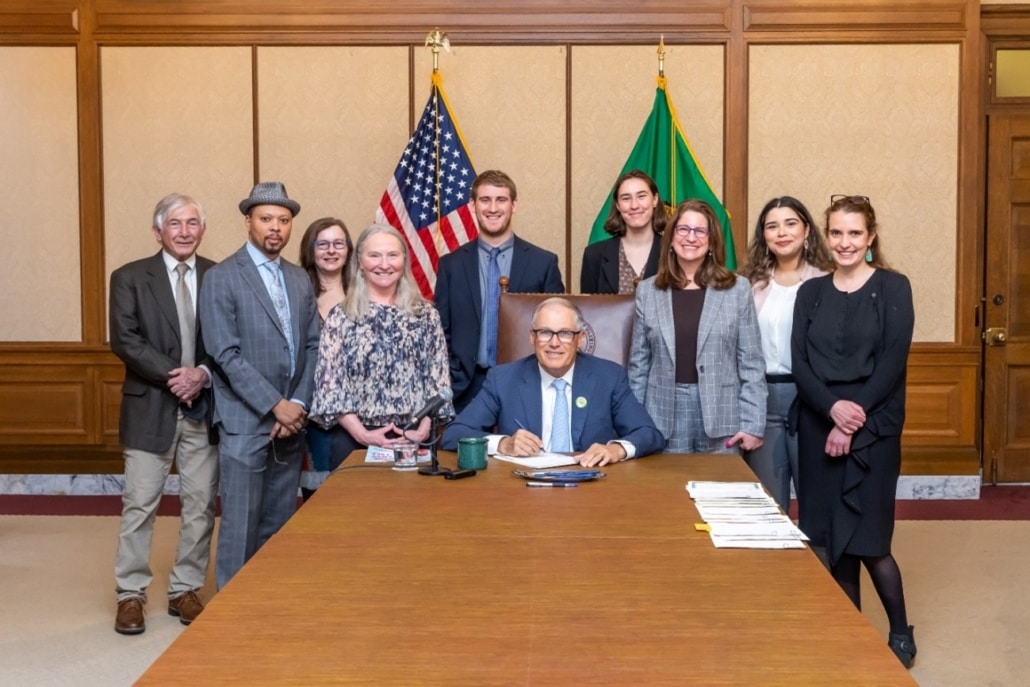 Supporters surround Governor Inslee as he signs ESHB 1394 into law. The U.S. and Washington State flags are in the background.