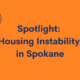An orange tile with dark blue text reads: Spotlight: Housing Instability in Spokane. There is a magnifying glass in the upper lefft corner and a house in the bottom right corner.
